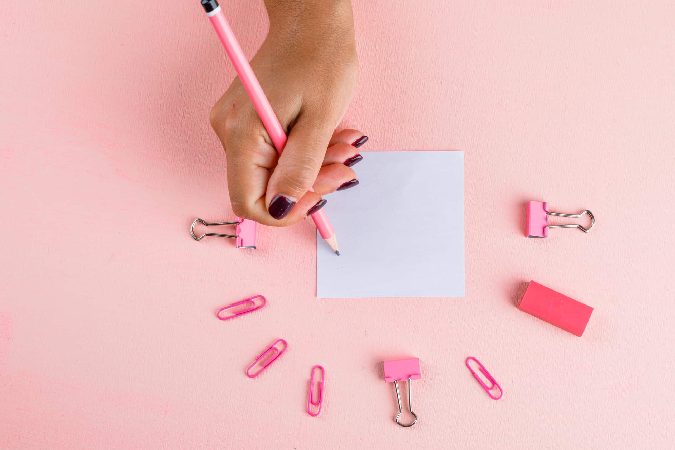 Mano escribiendo con un lapiz rosa sobre un papel cuadrado rodeado de clips y otros elementos de color rosa