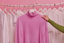 Mano que coge un jersey rosa de una percha con un fondo de camisas y camisetas colgas de sus perchas de colores blancos