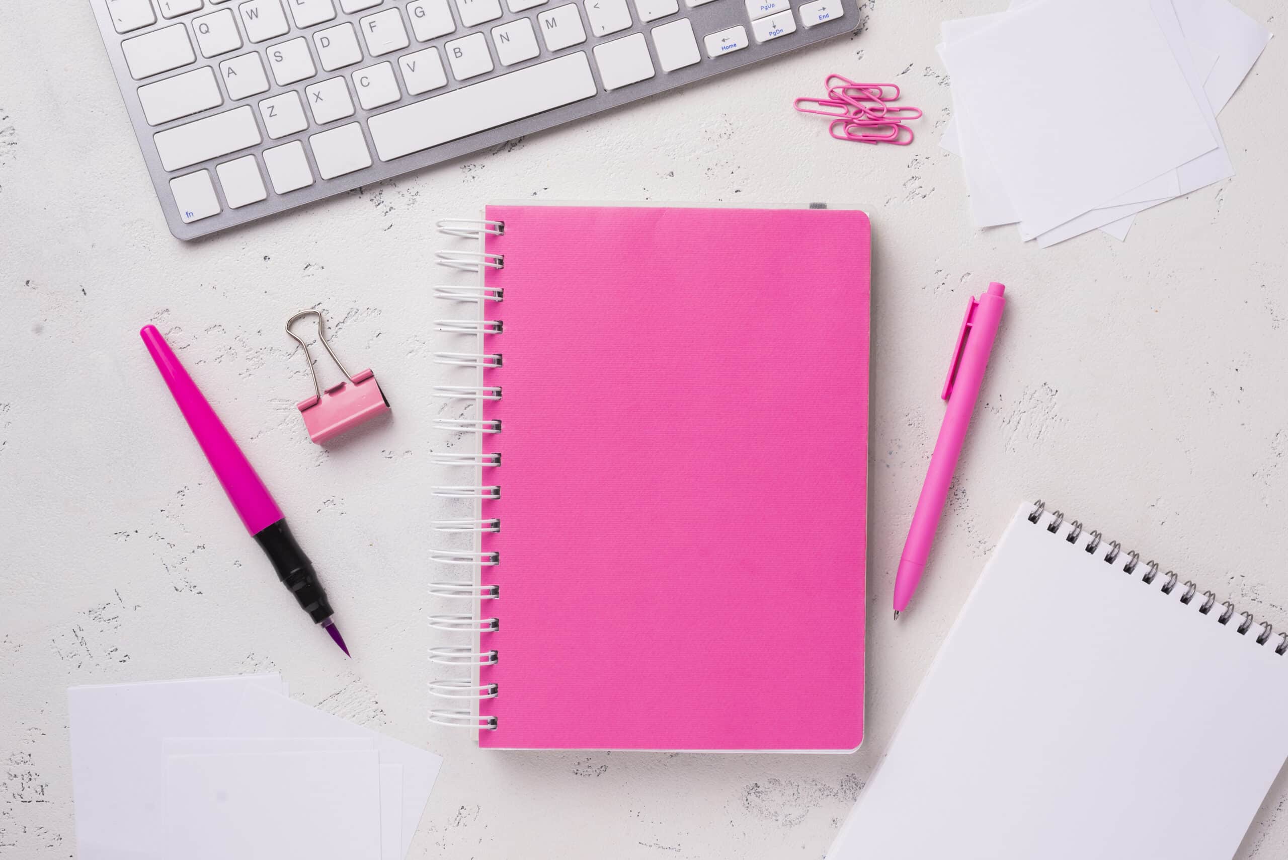 Una libreta rosa con elementos alrededor como boligrafos, clips, otra libreta y un teclado